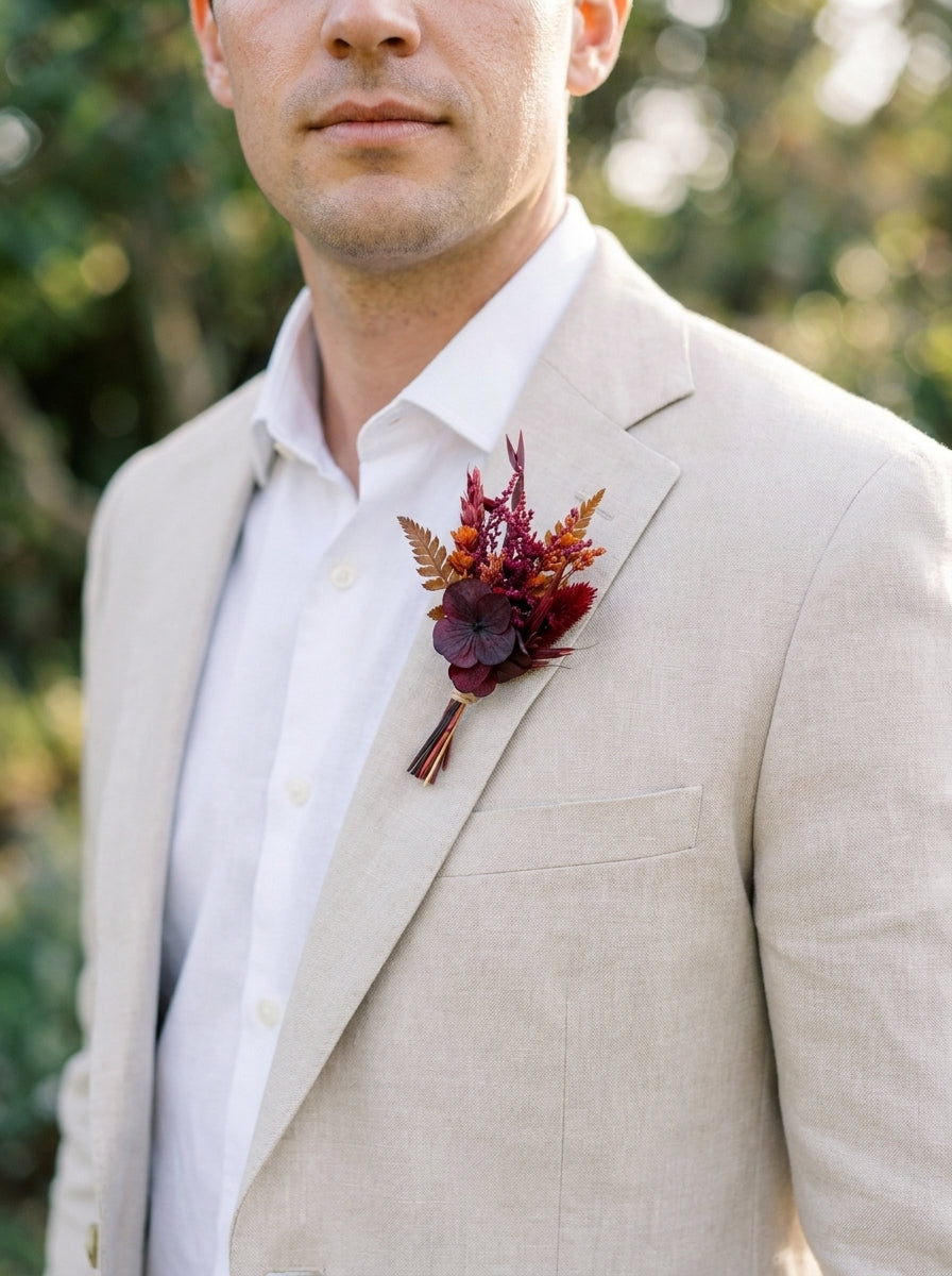 A man wearing a light beige linen suit jacket over a white dress shirt, featuring a rustic autumnal boutonniere with burgundy and rust tones on the chest, set against a soft, blurred green outdoor background.