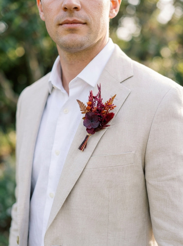 A man wearing a light beige linen suit jacket over a white dress shirt, featuring a rustic autumnal boutonniere with burgundy and rust tones on the chest, set against a soft, blurred green outdoor background.