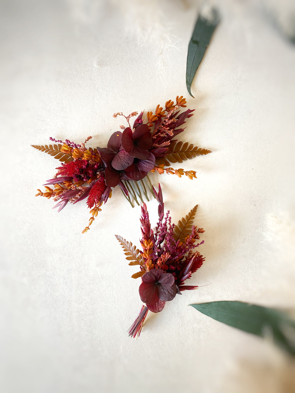 Hair comb and corsage with burgundy, rust orange, and brown botanicals arranged on a textured off-white surface.
