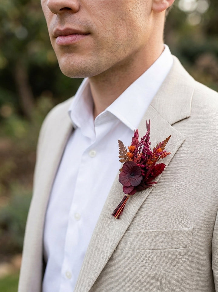 A close-up of a man wearing a light beige linen suit jacket, white dress shirt, and a dark red and orange dried floral boutonniere on his lapel, set against a soft, blurred outdoor background.