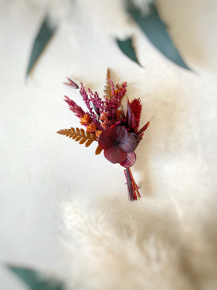 A small dried floral boutonniere with burgundy hydrangea, rust grasses, and brown ferns, photographed on a soft cream fabric background.