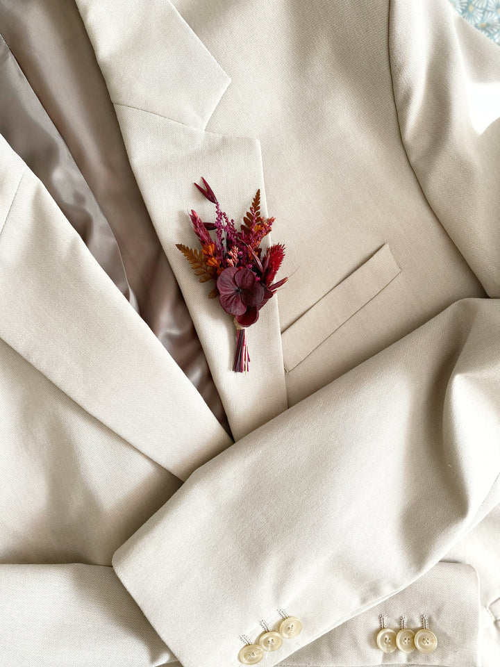 A close-up of a cream-colored suit jacket with a small dried floral boutonniere in burgundy, rust, and orange pinned to the chest pocket.