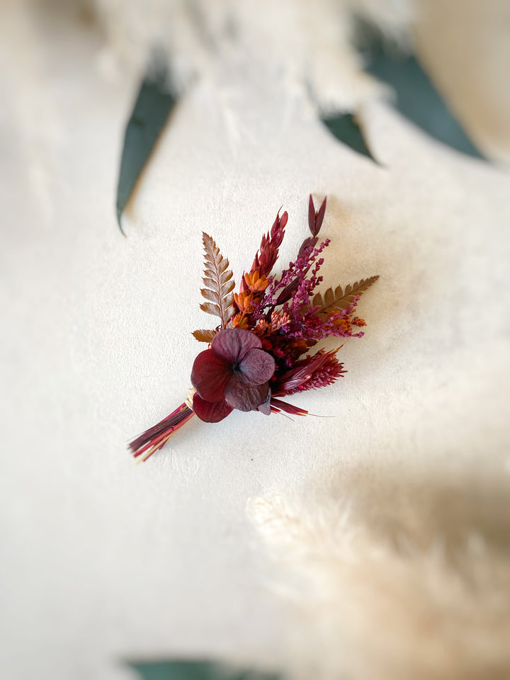 A small dried floral arrangement with burgundy hydrangea-like blooms, rust and orange filler stems, and muted green ferns, photographed on a soft off-white textured surface.
