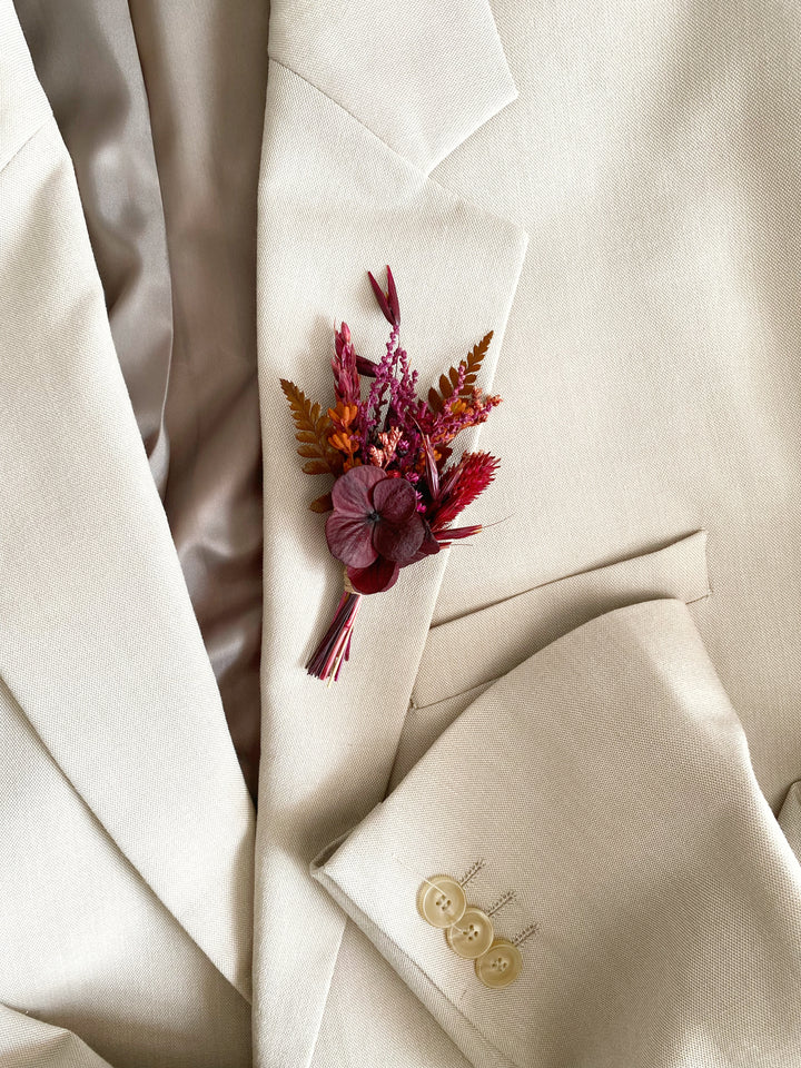 A close-up of a cream-colored suit jacket with a small dried floral boutonniere pinned to the chest pocket, featuring burgundy, rust, and orange botanicals.