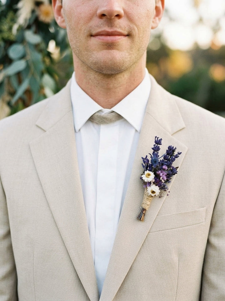 A close-up of a man wearing a light beige suit jacket, white dress shirt, and a beige tie, with a small floral boutonniere in purple and white tucked into his pocket. The background is a soft, blurred outdoor setting with green foliage.