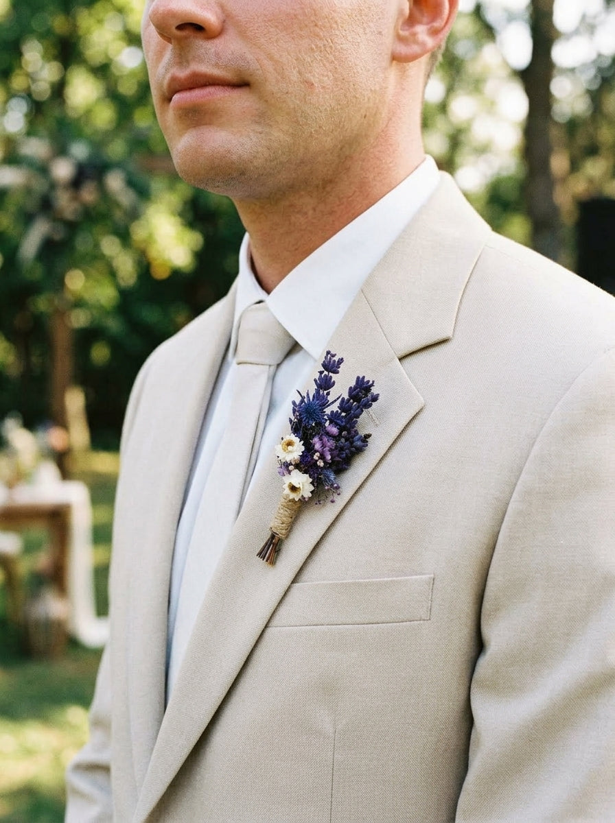 A close-up of a man wearing a light beige suit jacket, white dress shirt, and matching tie, with a purple and white floral boutonniere on the lapel, set against a blurred outdoor garden background.