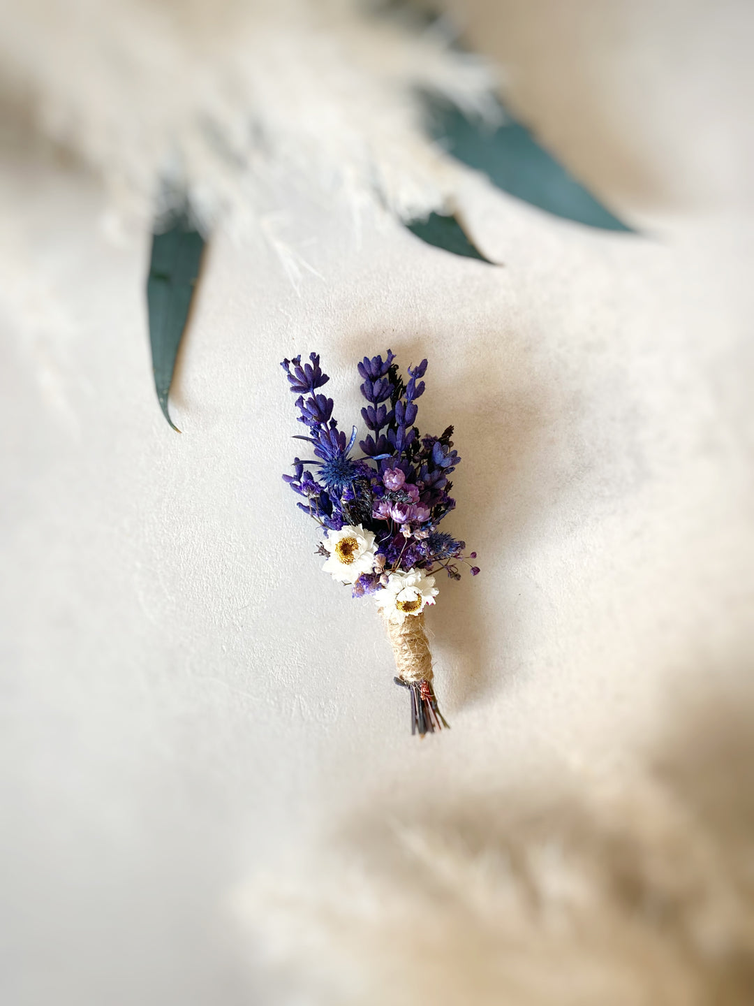A small dried floral boutonniere featuring purple lavender-like sprigs, tiny white daisy-like flowers, and a beige twine-wrapped stem, photographed on a soft cream surface with blurred green leaves in the background.