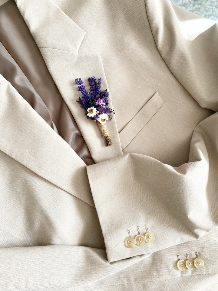 A close-up of a light beige or cream-colored suit jacket with a small floral boutonniere pinned to the chest pocket. The boutonniere features purple lavender-like stems and small white daisy accents.