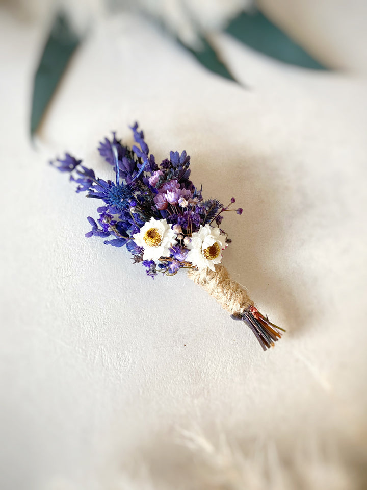 A small dried floral boutonniere featuring purple lavender, white daisies, and yellow centers, wrapped in natural twine, resting on a textured off-white surface.