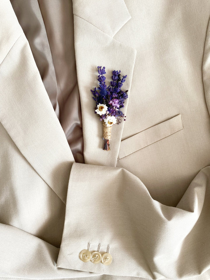 A close-up of a beige suit jacket with a small dried flower boutonniere pinned to the chest pocket, featuring purple lavender and white daisy elements.