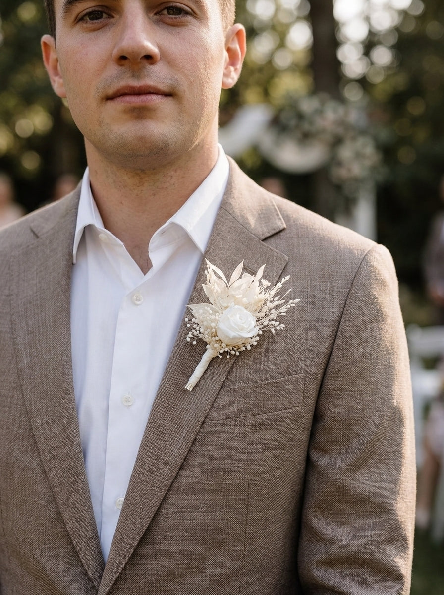 A man wearing a taupe-brown linen suit jacket over a white dress shirt, featuring a white floral boutonniere on the lapel, set against a soft-focus outdoor background.