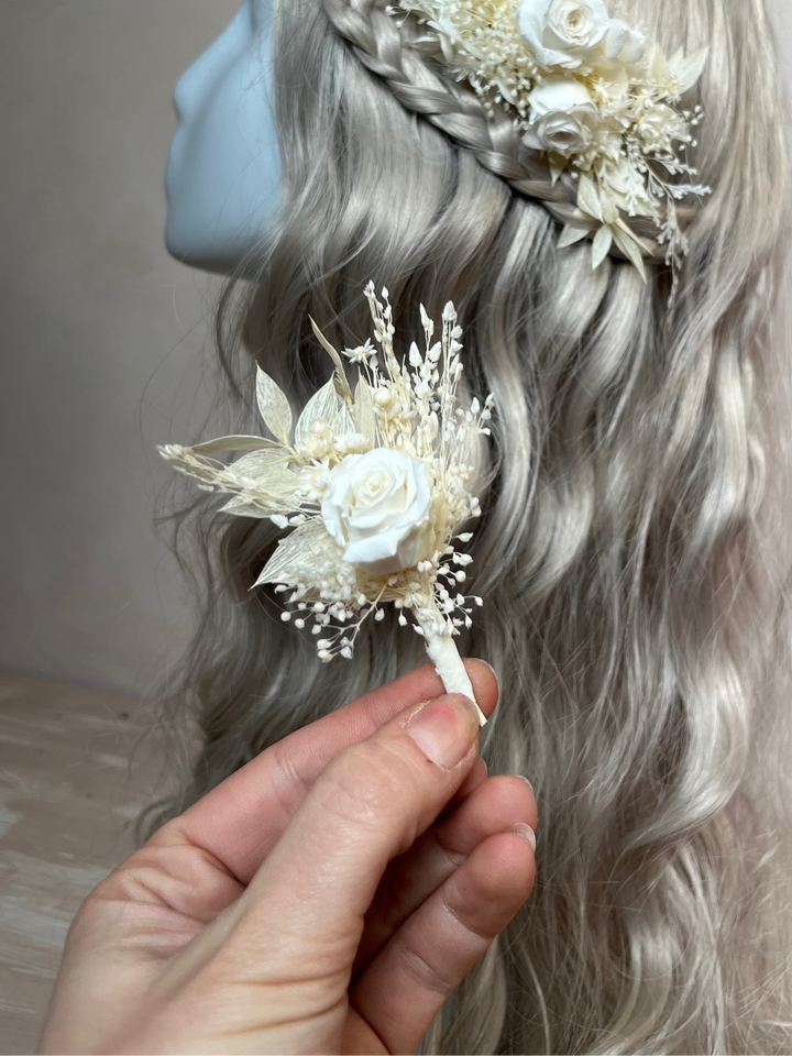 A hand holds a small cream floral boutonniere near a mannequin head with long wavy silver-gray hair, showing a matching braided hair piece and matching corsage in an off-white dried flower style.