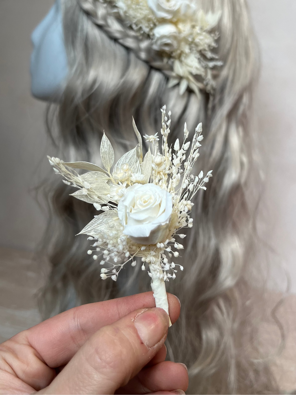 A hand holds a small dried floral boutonniere  in front of a mannequin head with long wavy light gray hair. The accessory features a white rose surrounded by cream and off-white dried leaves and tiny sprigs.
