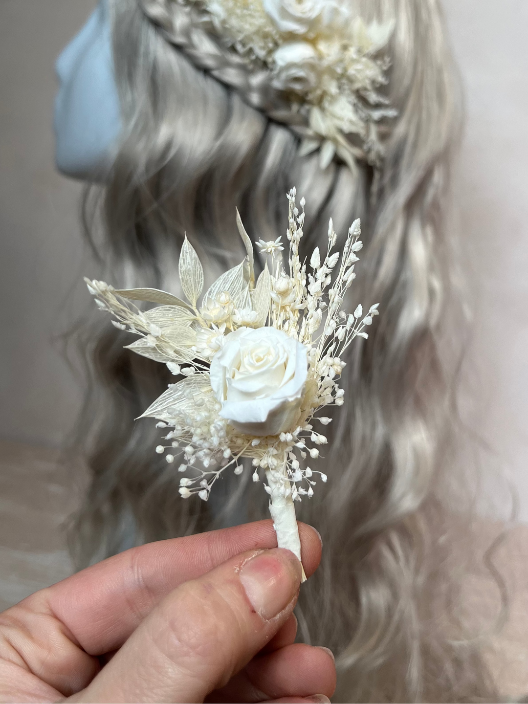 A hand holds a small dried floral boutonniere  in front of a mannequin head with long wavy light gray hair. The accessory features a white rose surrounded by cream and off-white dried leaves and tiny sprigs.