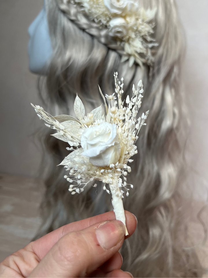 A hand holds a small dried floral boutonniere with cream and ivory flowers and delicate stems in front of a mannequin head with long wavy silver-gray hair wearing a larger matching floral headpiece.