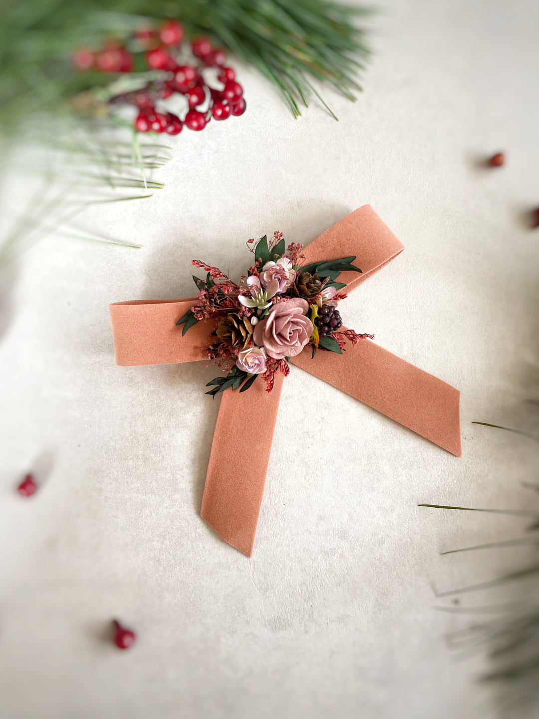 A floral hair bow with a peach ribbon bow, featuring pink and burgundy flowers with green foliage, styled on a textured cream surface with pine branches and red berries.