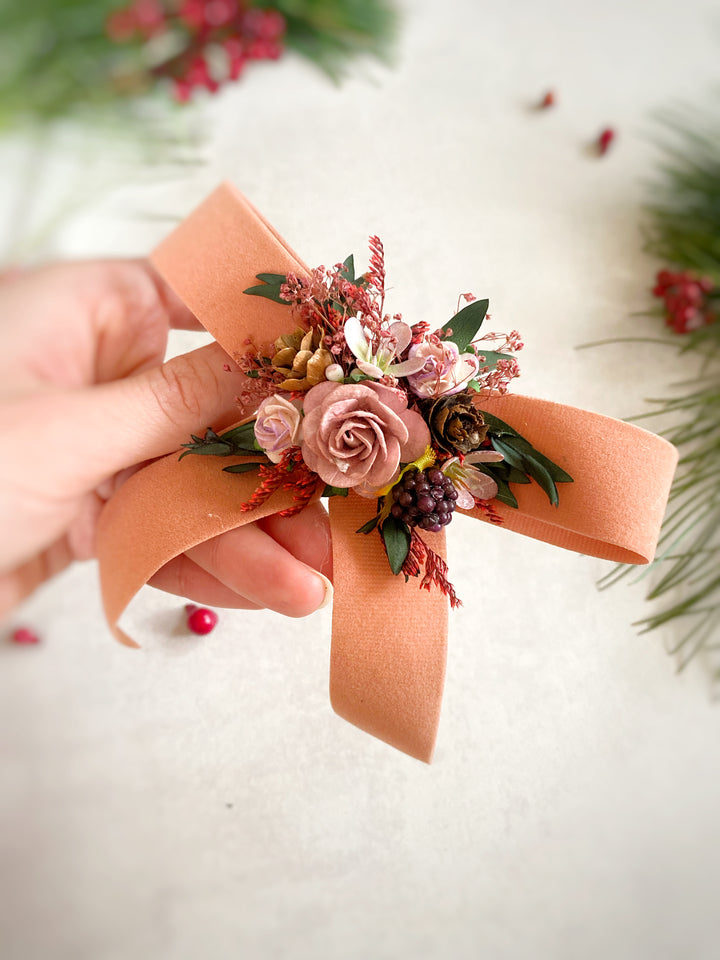 A hand holds a small floral hair bow or boutonniere-style accessory with a peach ribbon bow, pink roses, green leaves, and red berries against a soft light background with blurred greenery and red berries.