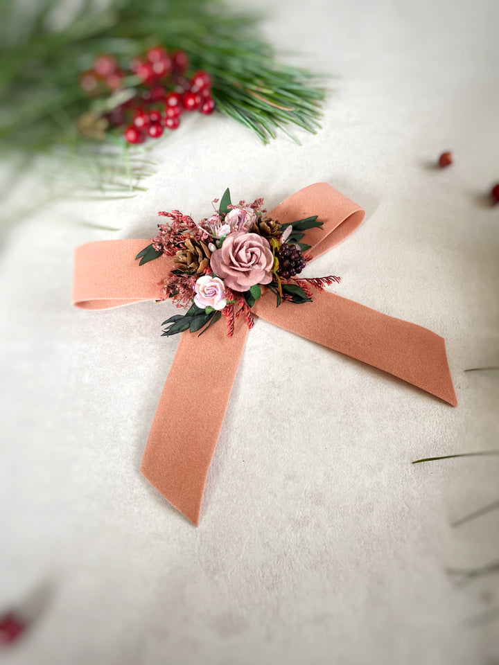 A peach-colored felt ribbon bow decorated with a small floral arrangement featuring pink and white roses, green leaves, and red berries, displayed on a textured off-white surface with blurred pine branches and red berries in the background.