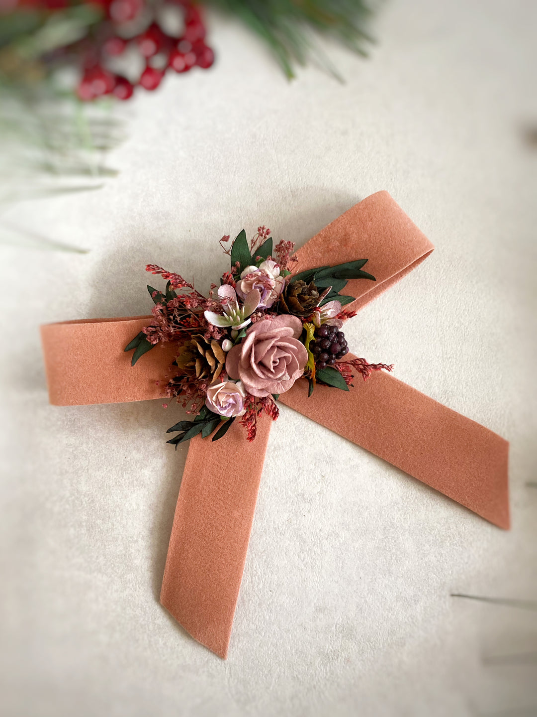 A soft orange-colored fabric bow with a floral boutonniere center, displayed on a textured off-white surface with a soft-focus holiday background.