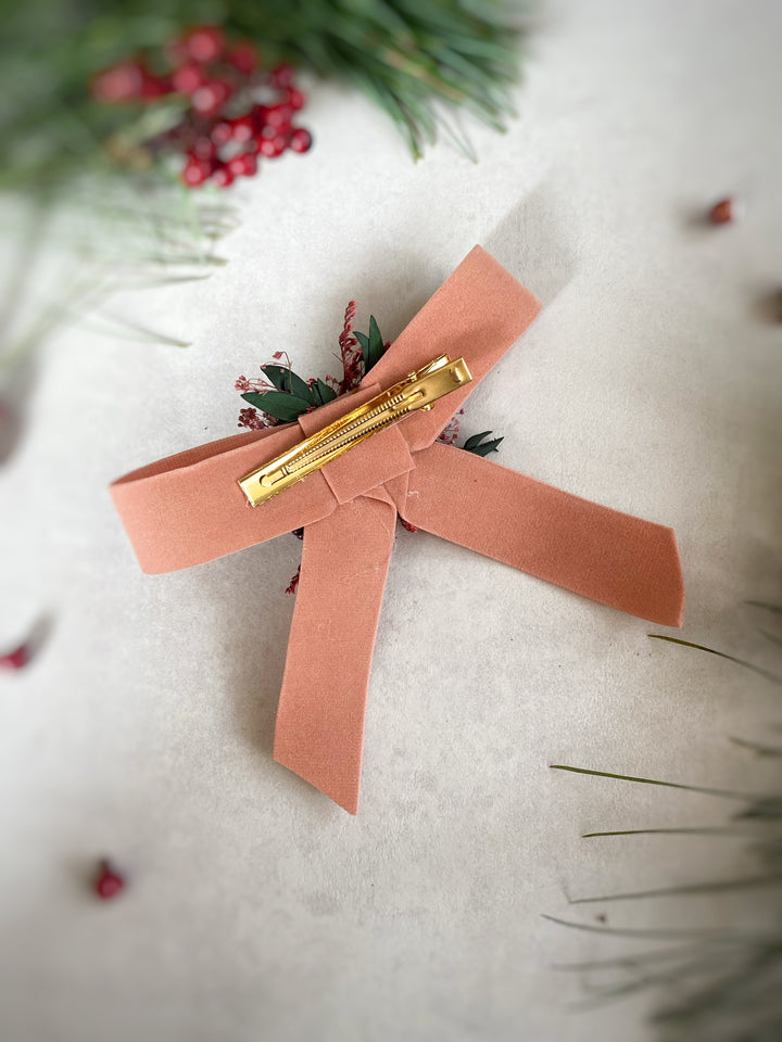A soft orange-colored felt hair bow with a gold metal claw clip, styled with small green pine leaves and red berries on a textured light beige background.