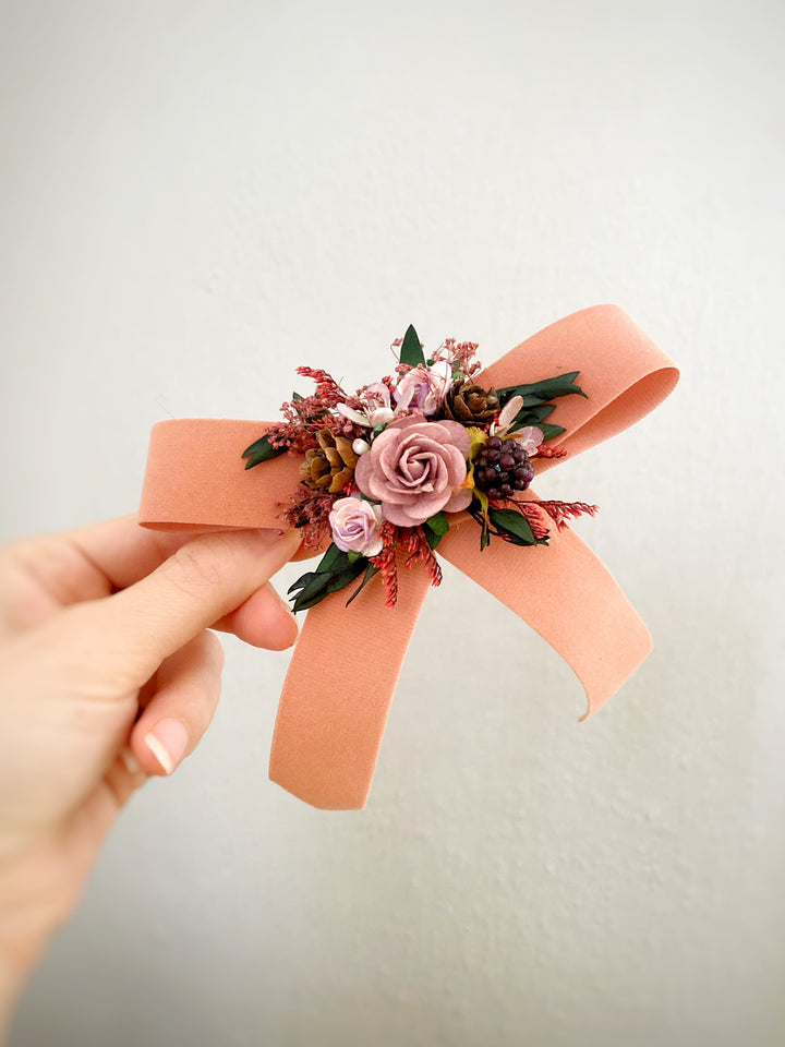 A peach-colored fabric bow headband with a central floral embellishment, held by a hand against a plain light gray wall.