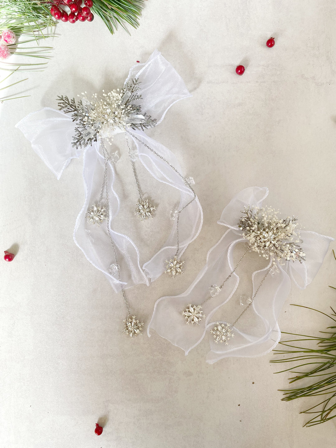 Two white tulle Christmas hair bows with silver and white beadwork, displayed on a light beige textured background with holiday greenery and red berries.