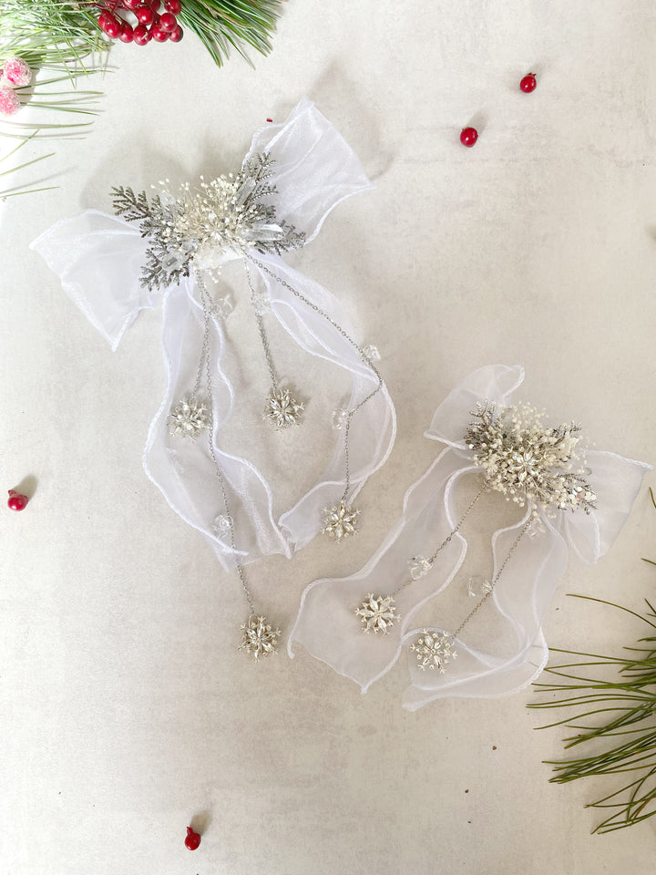 Two white tulle Christmas hair bows with silver and white beadwork, displayed on a light beige textured background with holiday greenery and red berries.