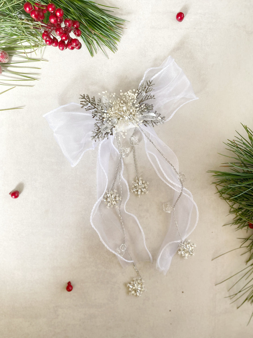 A decorative Christmas  bow accessory made of sheer white organza ribbon, silver glittery pine branches, and white bead snowflake charms, arranged on a beige surface with holiday greenery and red berries as props.
