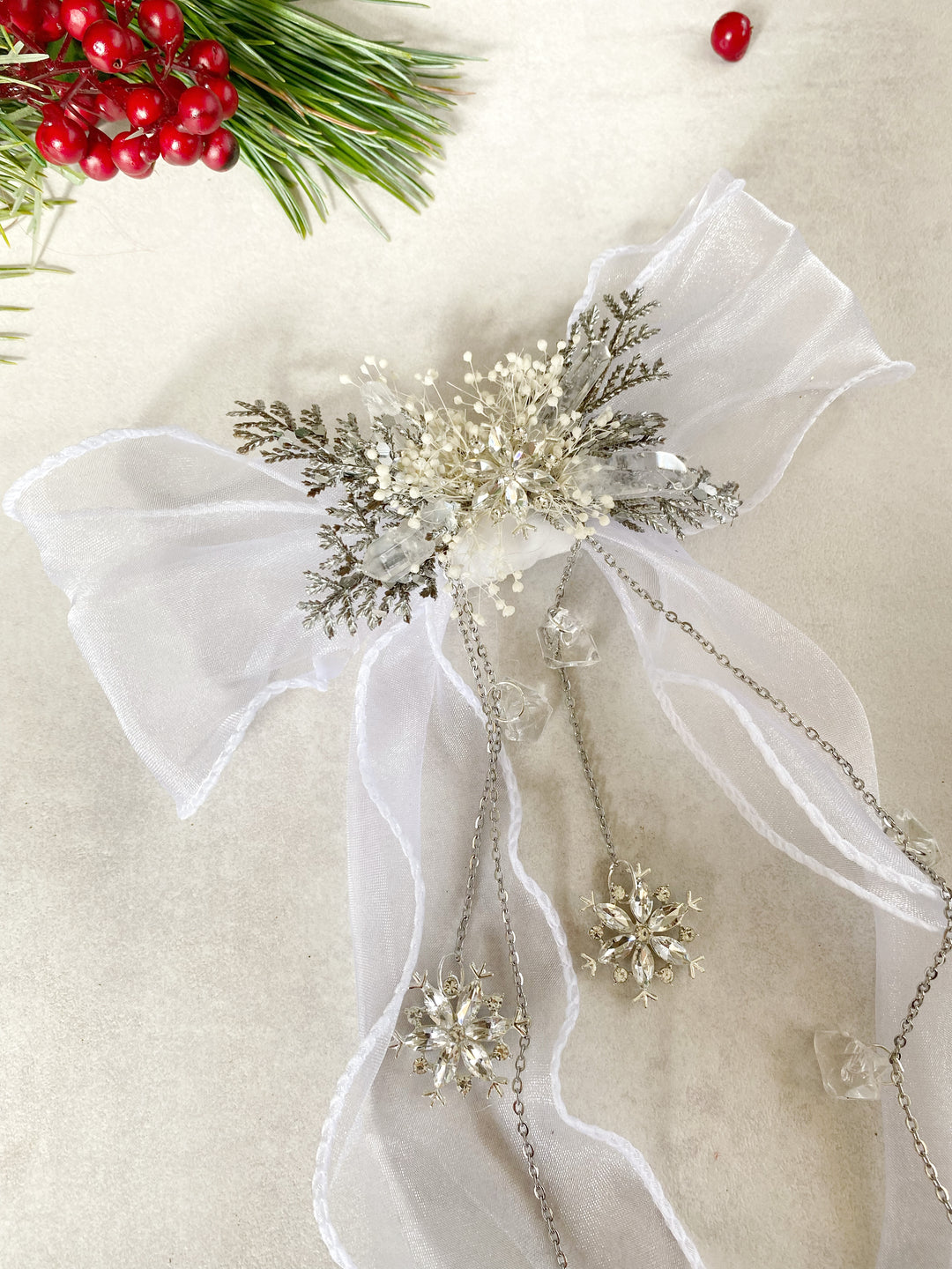 A white organza bow decorated with a central silver and white snowflake embellishment, delicate chains, and small clear crystals, styled on a light beige surface with holiday greenery and red berries in the corner.