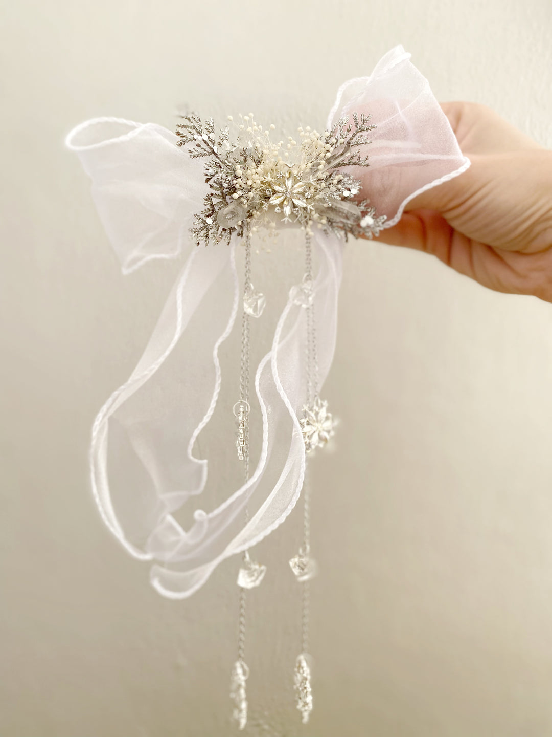 A hand holding a white organza bow hair accessory decorated with a central cluster of silver and gold glittery snowflakes, clear rhinestones, and dangling clear bead chains.