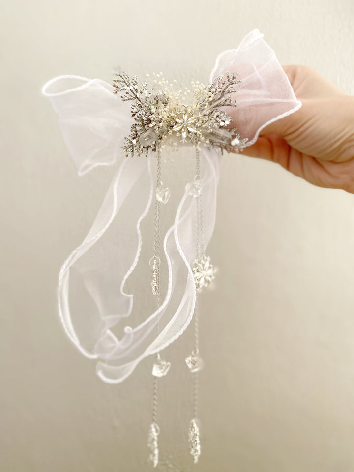 A hand holding a white organza bow hair accessory decorated with a central cluster of silver and gold glittery snowflakes, clear rhinestones, and dangling clear bead chains.