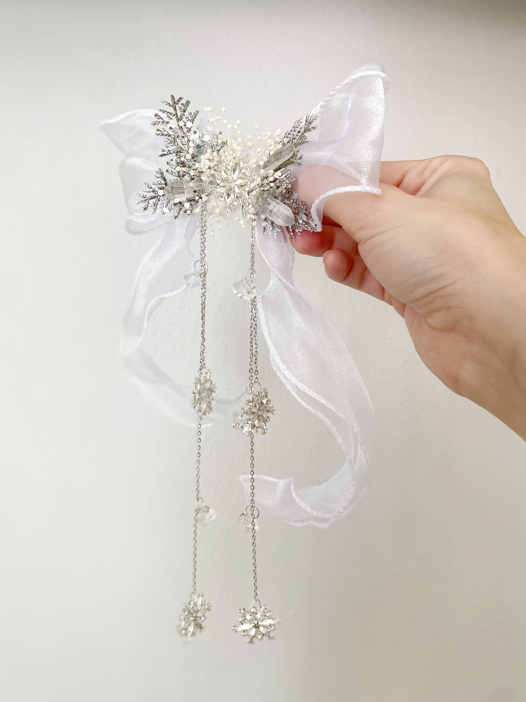 A hand holding a white organza bow hair accessory embellished with silver glitter, rhinestones, and dangling snowflake charms, set against a plain light background.