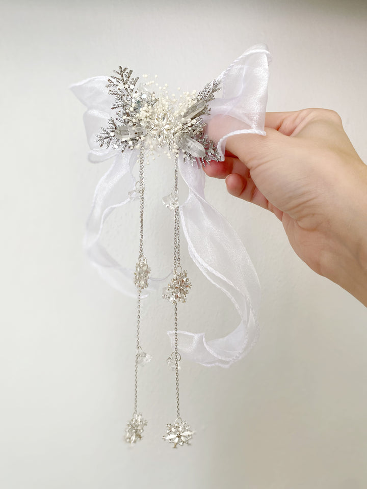 A hand holding a white organza bow hair accessory embellished with silver glitter, rhinestones, and dangling snowflake charms, set against a plain light background.