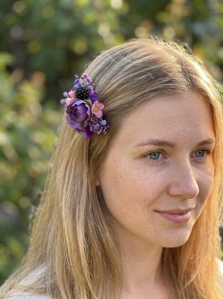 A close-up portrait of a woman with blonde hair wearing a small purple and pink floral hair accessory clipped to the side of her hair, set against a soft, blurred green outdoor background.