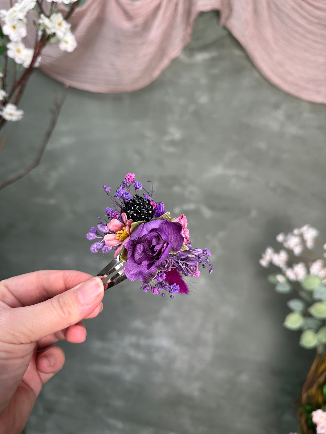 A hand holds a small floral hair accessory featuring a prominent purple rose, pink blossoms, a blackberry, and tiny purple filler flowers against a soft gray backdrop with blurred decorative flowers.
