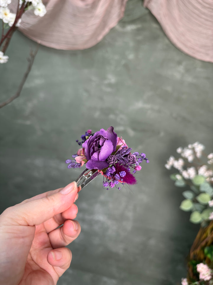 A hand holding a small purple floral hair clip with textile and preserved purple flowers against a soft gray-green textured background with blurred decorative flowers.