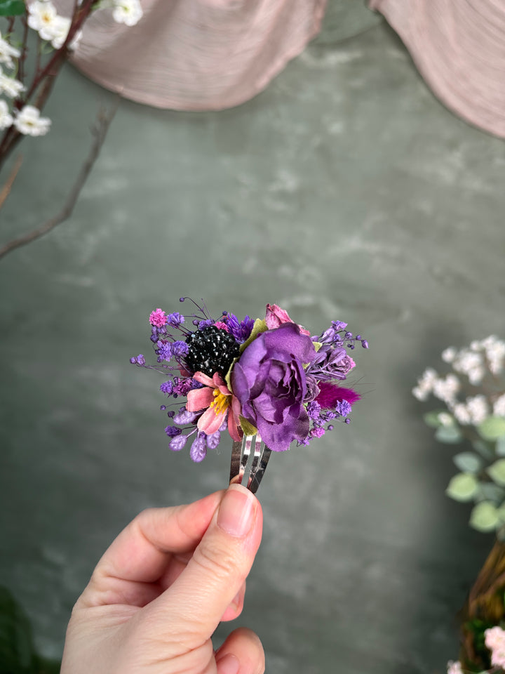 A hand holding a small floral hair accessory featuring a purple rose, purple wildflowers, and a black berry-like accent on a clear stand, set against a soft gray-green textured background with blurred floral elements.