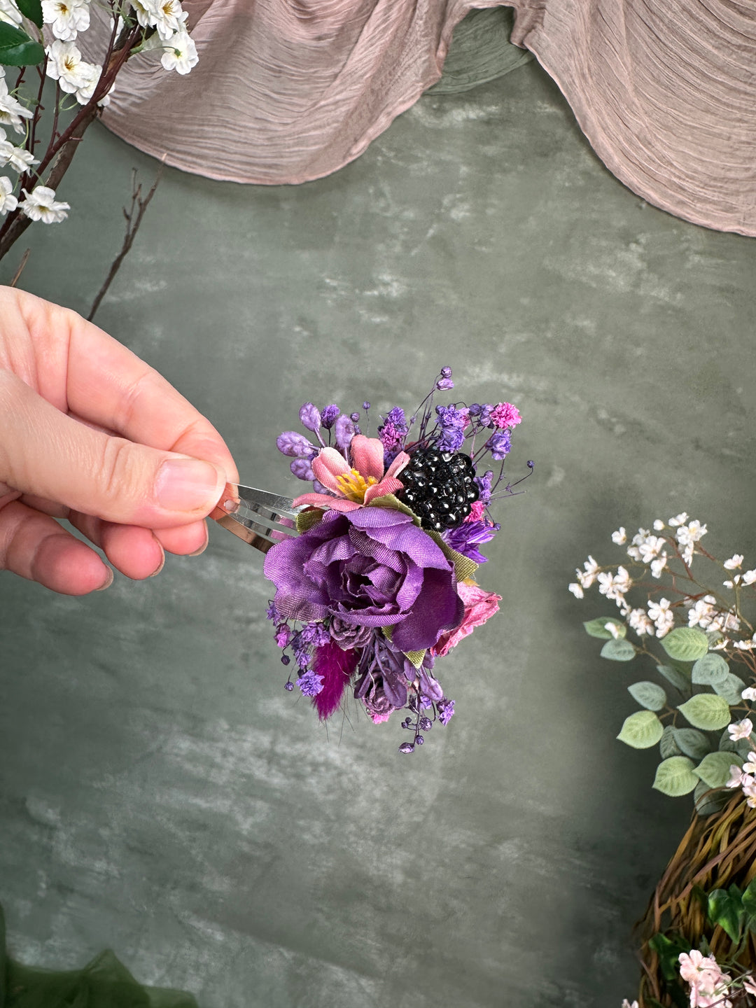 A hand holds a small floral hair clip decorated with purple and pink flowers, black berries, and tiny purple blossoms, set against a textured gray background with decorative fabric and flowers.