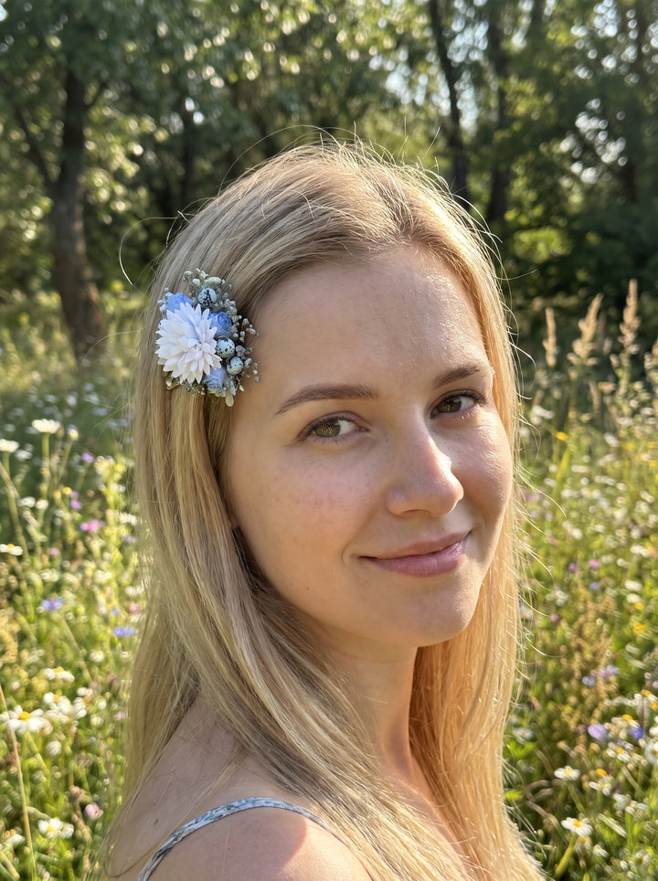 A woman with long blonde hair wearing a decorative hair accessory with  light blue flowers, photographed outdoors in a sunlit grassy field.