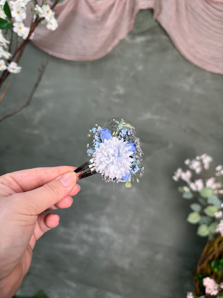 A hand holds a small floral hair clip featuring  light blue flower with tiny green leaves and clear accents, set against a soft gray textured background with blurred decorative flowers and draped fabric.