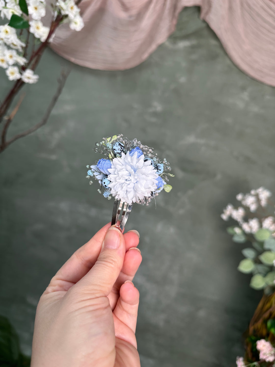 A hand holds a small floral hair clip featuring a  light blue flowers and blueberries, set against a textured gray-green background with decorative flowers and draped fabric.