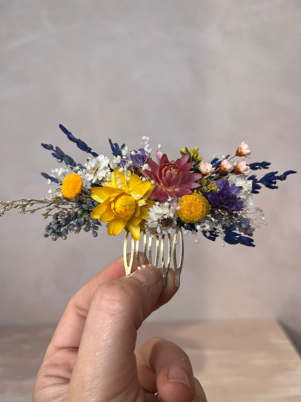 A hand holding a gold-toned hair comb decorated with a small dried floral arrangement featuring yellow, purple, pink, and white flowers against a neutral gray background.