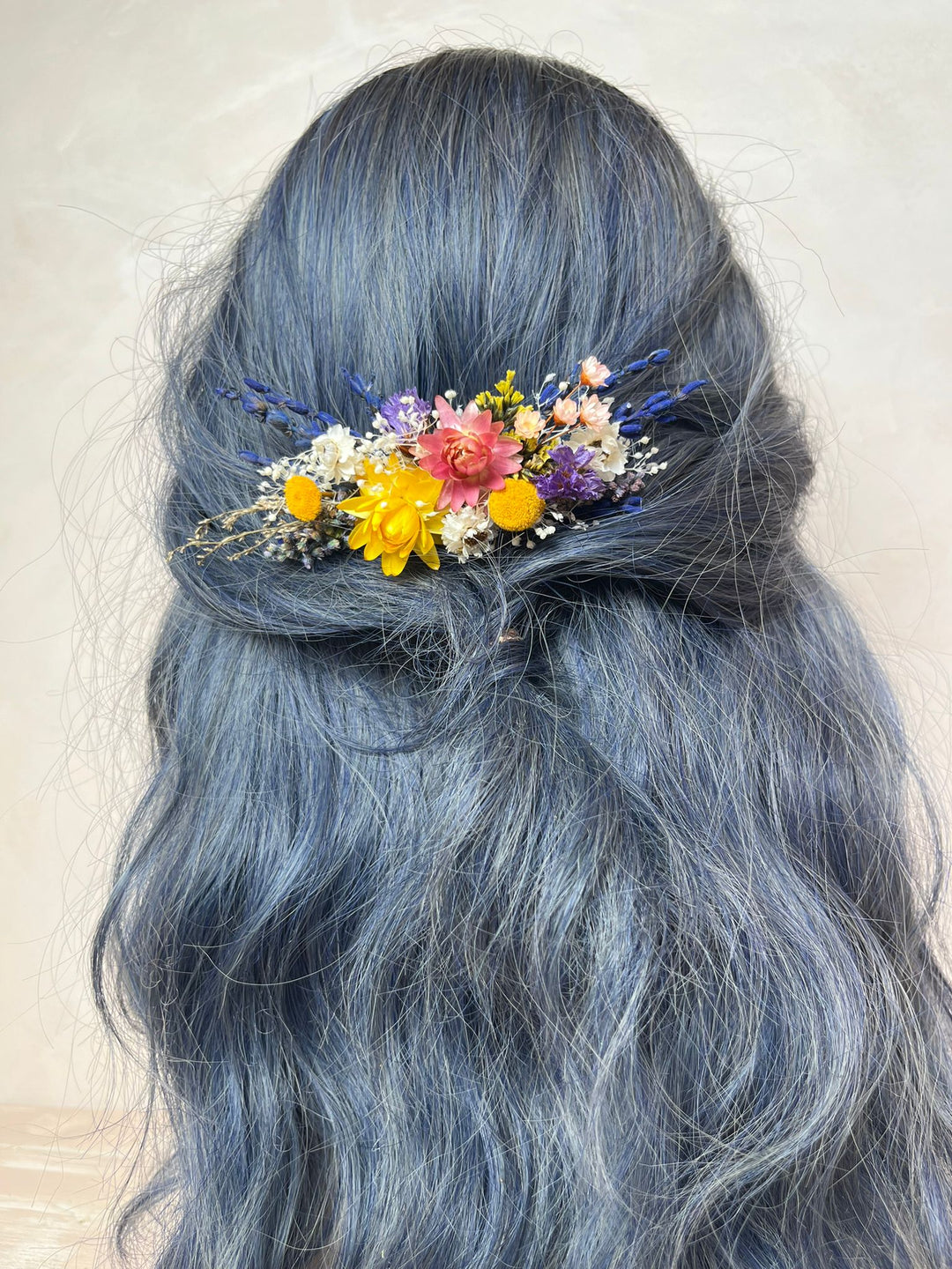A close-up of a person's long, wavy, slate-blue hair styled with a small, colorful dried flower hair accessory at the back of the head.