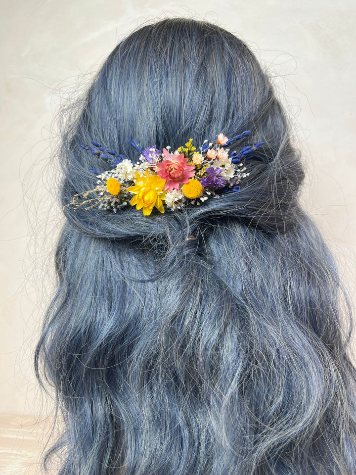 A close-up of a person's long, wavy, slate-blue hair styled with a small, colorful dried flower hair accessory at the back of the head.
