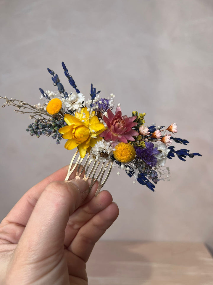 A hand holding a small dried flower hair comb with yellow, pink, purple, and blue blossoms against a soft neutral background.