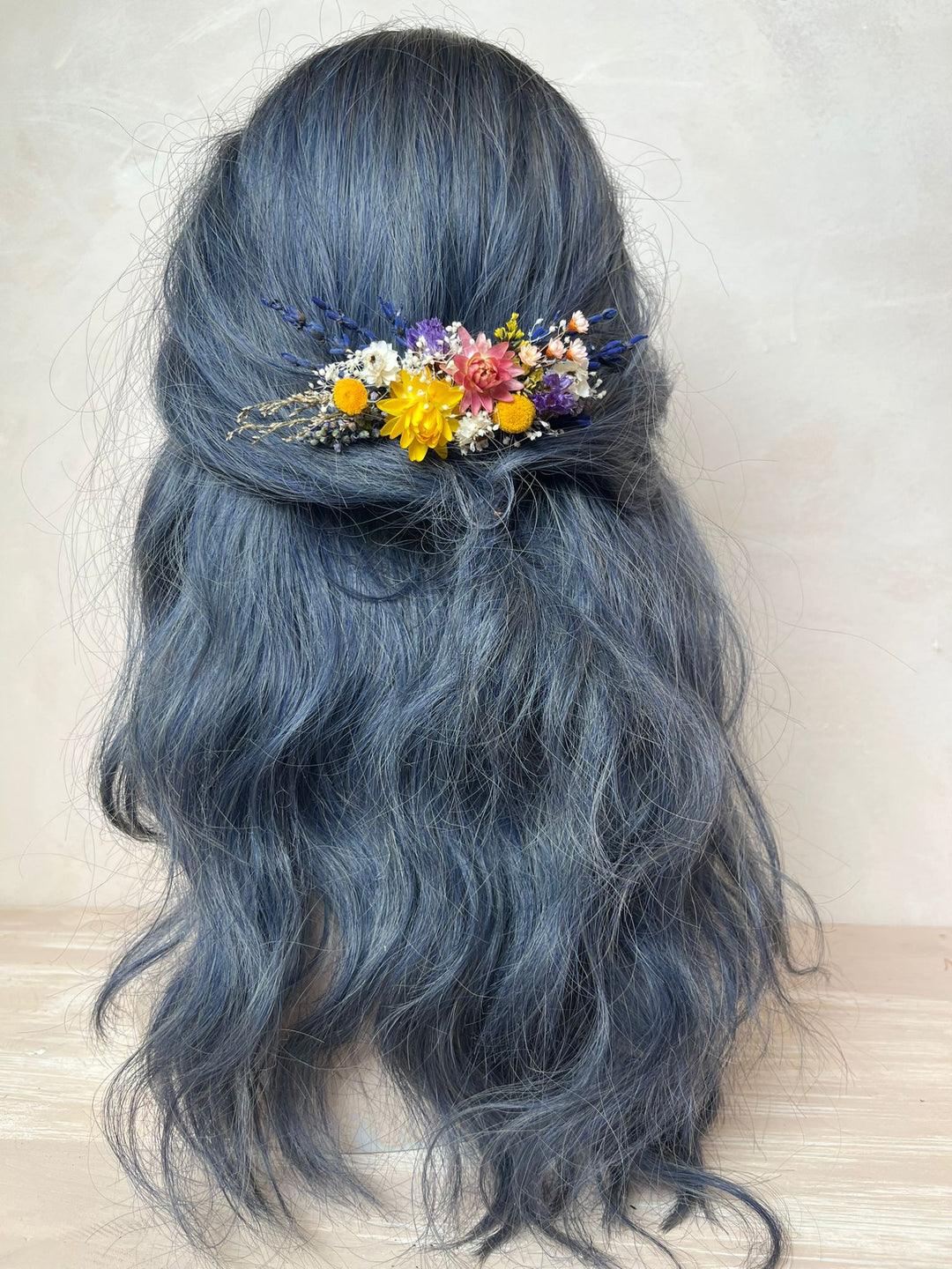 Long wavy dark blue-gray hair styled in a half-updo with a small multicolor dried flower hair accessory, shown on a mannequin head against a neutral beige background.