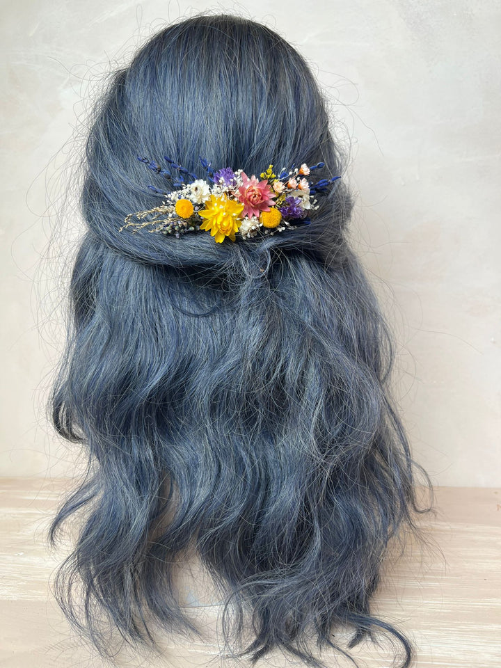 Long wavy dark blue-gray hair styled in a half-updo with a small multicolor dried flower hair accessory, shown on a mannequin head against a neutral beige background.