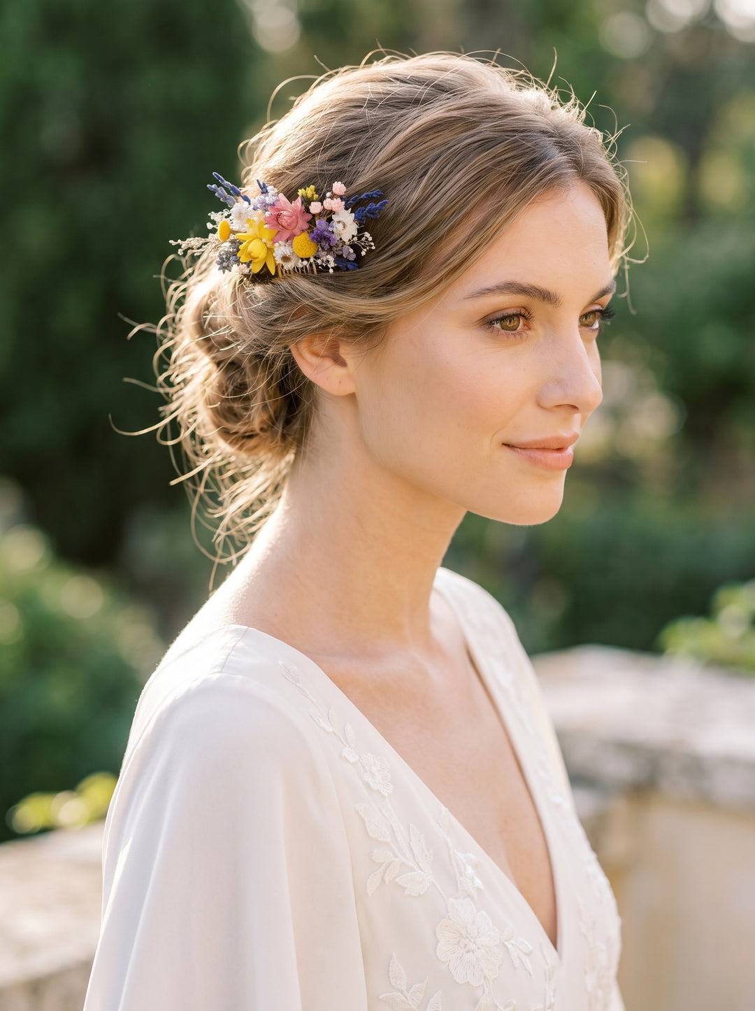A woman in a white lace wedding dress wearing a delicate multicolor floral hair accessory with small blue and yellow flowers, photographed outdoors with a soft blurred green background.