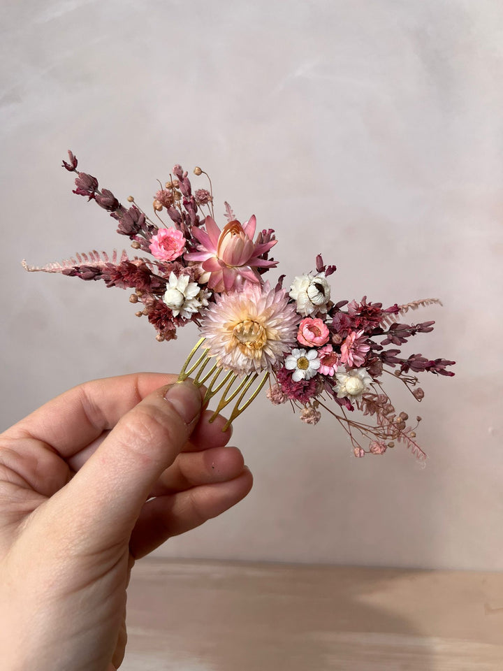 A hand holding a gold-toned hair comb decorated with dried pink, burgundy, and white flowers against a soft beige background.