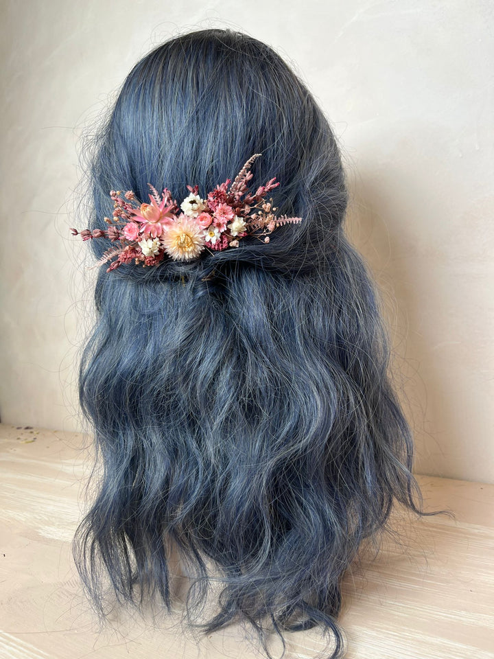 A long, wavy dark gray wig with a floral hair accessory featuring pink, orange, and cream dried flowers, shown from the back against a neutral beige background.
